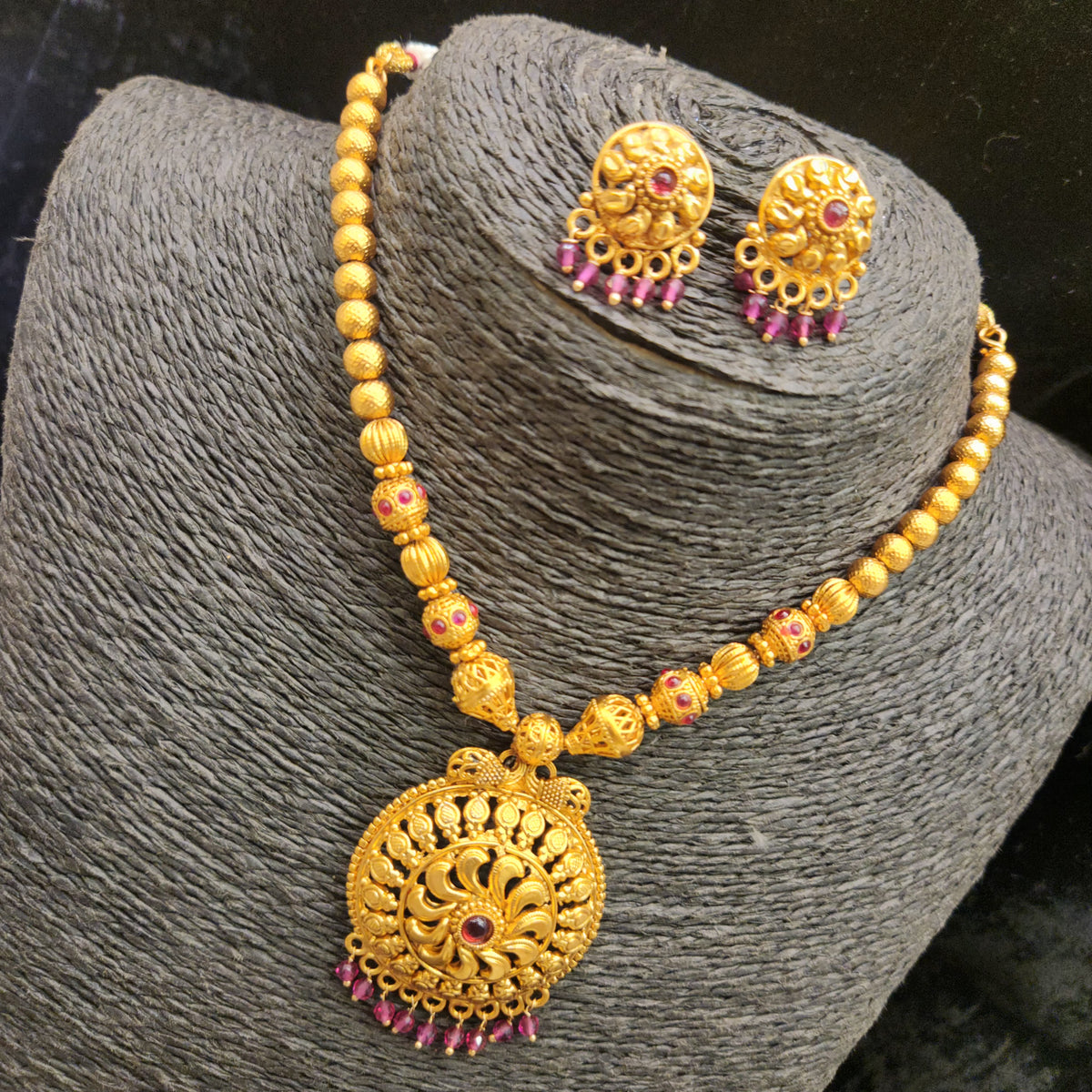 Gold-toned traditional necklace and matching stud earrings displayed on a dark gray textured jewelry bust. The necklace features a large ornate round pendant with floral filigree and small pink gemstone accents, along with matching round stud earrings.